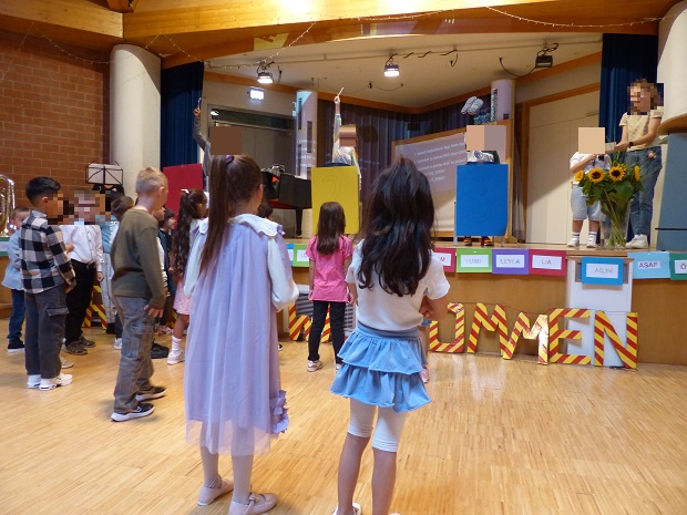 Kinder stehen vor der Bühne, auf der 3 weitere Kinder stehen und Gegenstände hochhalten für das Spiel 1, 2 oder 3 © Nora Leiner